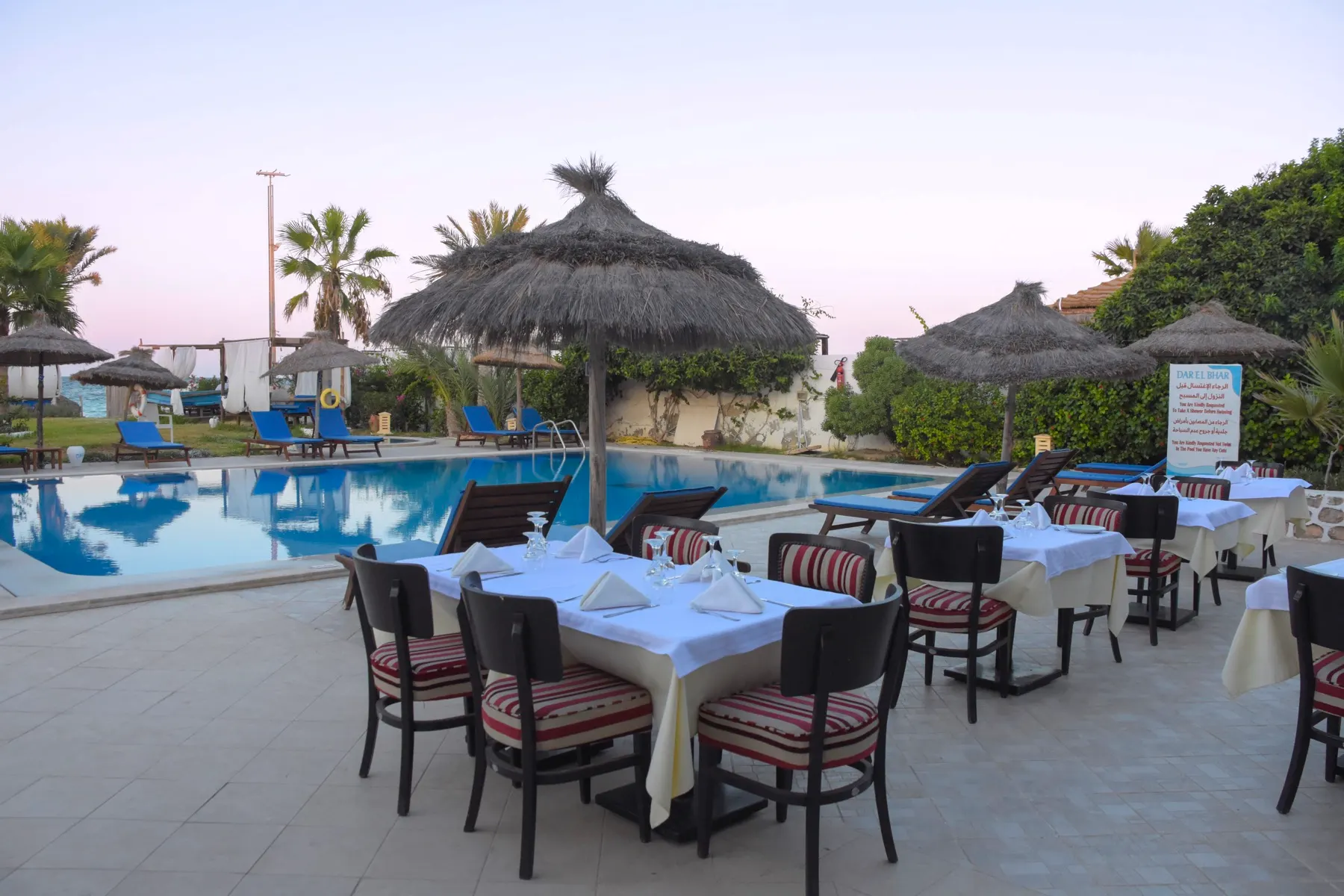 Table dressée au bord de la piscine de Dar El Bhar au coucher du soleil, sous les parasols en palmes