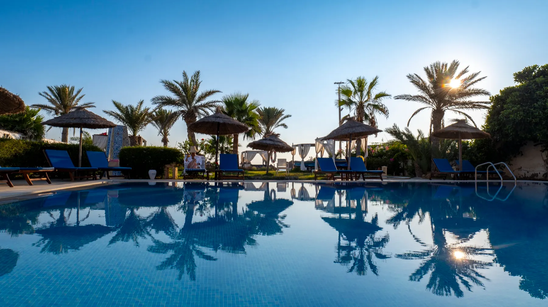 La piscine de Dar El Bhar à l'aube, palmiers et parasols en palmes reflétés dans l'eau calme